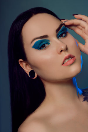 Brunette girl with tunnels in her ears in a beautiful blue light. Woman with blue make-up, fashionable blue arrows over the eyelid. Insightful brown eyes. Model with beautiful hair and professional makeup.の写真素材