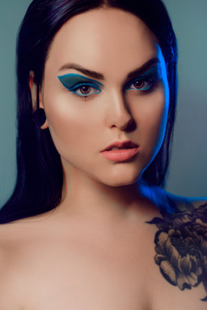 Brunette girl with a flower tattoo on her shoulder and tunnels in her ears in beautiful blue light. Woman with blue make-up, fashionable blue arrows over the eyelid.の写真素材