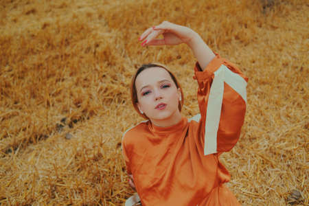 A gorgeous girl in a long dress sits on a wheat field. Blonde posing for a fashionable photo. Woman moves her hands beautifully.の写真素材