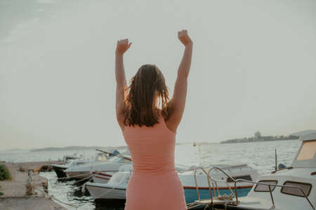 A joyful girl against the background of the sea raised her hands up. Vacation, freedom. Woman on the background of yachts and shipsの写真素材