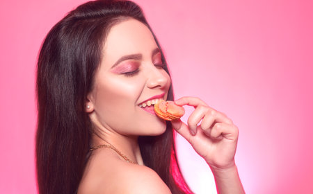 Girl eats macaroon, model on a pink background eats a sweet, sugar diet. Brunette with pink make-up, addicted to food. Macaroon bites, cookiesの写真素材