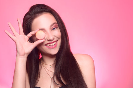 Girl eats macaroon, model on a pink background eats a sweet, sugar diet. Brunette with pink make-up, addicted to food.の写真素材