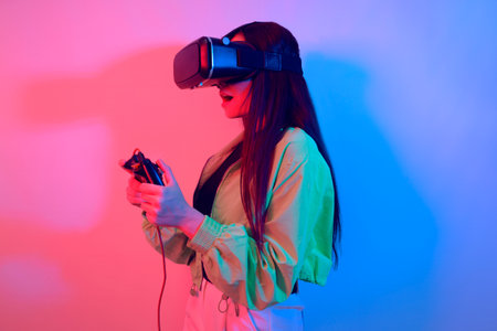 A shocked girl in neon with VR glasses holds a gamepad in her hands and plays games. Virtual world, technology.の写真素材