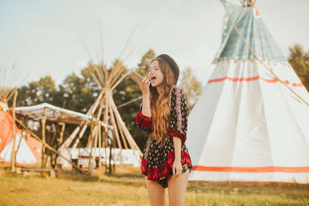 Young girl beautiful tourist talking on the phone, video communication roaming on the background teepee / tipi- native indian house. Unusual places to stayの写真素材