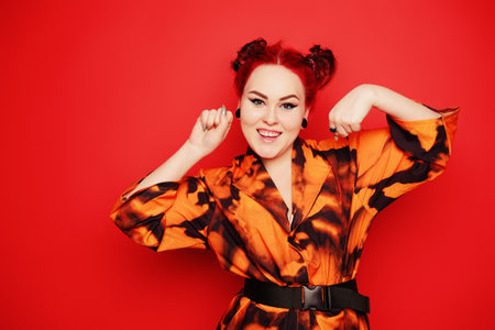 Cute girl on a red background. Red hair and tunnels in the ears, red dress and lips. Anime poses. The girl with eyeliner and two buns on her head grimaces, smiles, has fun. Japanese cultureの写真素材