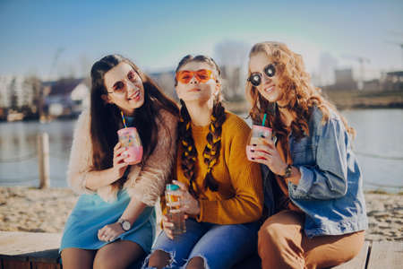 Three girls sit on the beach, near the river, talk, gossip, joke, laugh, smile at womans day. Girls...