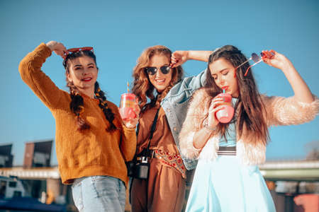 Three girls near the river against the sky having fun. Fashion girls in sunglasses. Jump, rejoice, drink drinks, smile have fun, going crazy. Color dressed friends spend summer vibes, danceの写真素材