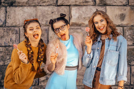Beautiful teenage girlfriends in trendy dresses and glasses walk in the summer. Sunny day, summer mood, fun and happiness. Three girls are smiling and eating a lollipop.の写真素材