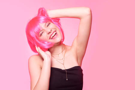 The girl in a pink wig, on a pink background, is happy, grimaces, smiles. Bright photo of a...
