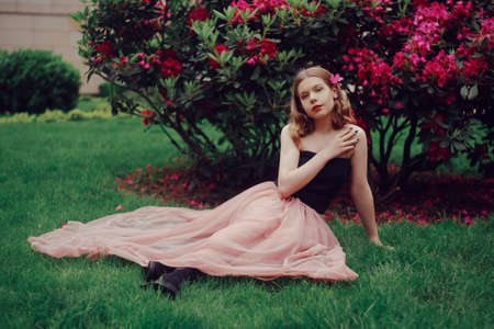 A girl in a long pink dress sits in the garden on the grass against a background of pink flowers. Short-haired blonde, fashionable princess. Flowers in her hair, looking at the camera, smiling.の写真素材