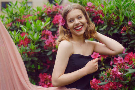 A girl in a long pink dress sits in the garden on the grass against a background of pink flowers. Short-haired blonde, fashionable princess. Flowers in her hair, looking at the camera.の写真素材