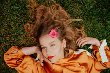 A girl with healthy beautiful hair lies on the grass and looks at the camera. Girl with perfect skin and natural care. Flower in the mouth holds a pink flower near the face. Natural beauty, youth, teenager. Red lipsの写真素材