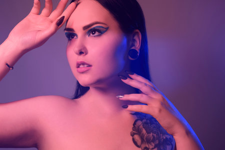 Brunette girl with a flower tattoo on her shoulder and tunnels in her ears in beautiful blue light. Woman with purple make-up, fashionable blue arrows over the eyelid.の写真素材