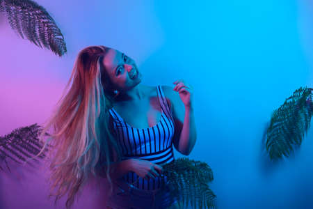Dance party summer vibe. A model in neon poses. Fashion photo of a girl in a swimsuit and shorts in pink-blue light. Girl on a background of tropical leaves summer photo.の写真素材