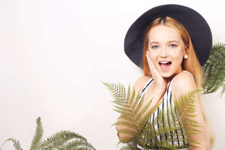 Girl in tropical leaves in a swimsuit in a big black hat smiles. Space for text, empty space point hand. Model summer photo. Big earringsの写真素材