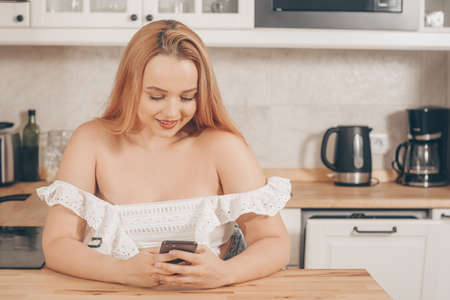 Beautiful woman sits at a table on the background of the kitchen at home. Girl with a smart phone in her hands and looks to the side pointing her finger to the side. Online delivery.の写真素材