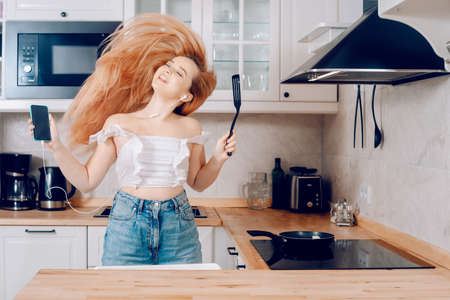 Woman singing and dancing in the kitchen with a smartphone, music. A girl in headphones with a phone stands in the kitchen and prepares food behind an induction stoveの写真素材