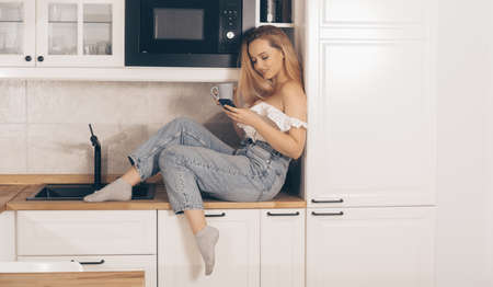 Food delivery, mobile shops, online shopping. A woman at home in the kitchen at the will of the refrigerator. A girl with a good figure holds a mobile phone in her handsの写真素材