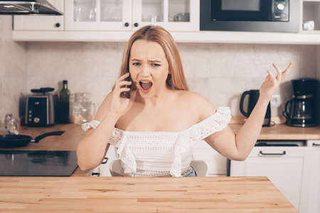 The woman looks into the laptop and rejoices, the girl sits at home against the background of the kitchen and smiles. The blonde won something online, found a job online.の写真素材