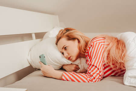 A girl looks at the phone in the morning, a woman in bed watches a video or reads the news. Internet addiction.の写真素材