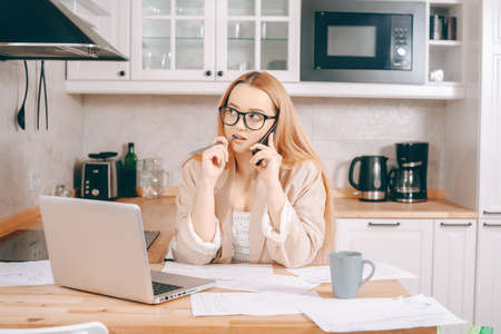 The woman at the laptop works from home in the call center. A beautiful blonde in a jacket and glasses looks into a laptop and writes something down. Girl speaks on the phone and processes the order.の写真素材