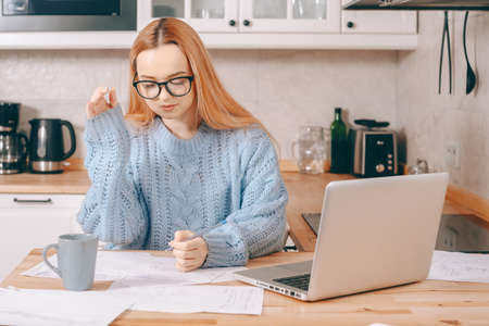A woman works from home using a laptop. A beautiful blonde in a sweater and glasses, there is a mug and papers on the table. Home schooling, work online. A student with headphones speaks by video callの写真素材