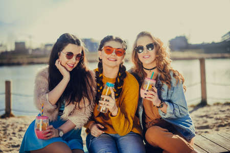 Three girls sit on the beach, near the river, talk, gossip, joke, laugh, smile at womans day. Girls on a hot day eat ice cream. Beauty model wearing sunglasses at summer day. Girl with hairslyleの写真素材