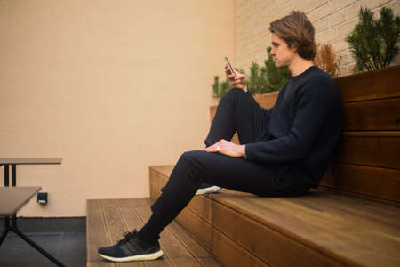 A portrait of a student guy sits on a bench and looks at the phone. A man looks at the screen and writes SMS, good weather. The guy in the sweaterの写真素材