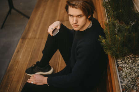 A man sits on a bench with a phone. Fashion man looks into the distance. Fashion man outside in sweater.の写真素材