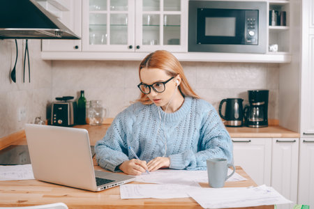 A woman works from home using a laptop. A beautiful blonde in a sweater and glasses, there is a mug and papers on the table. Home schooling, work online. A student with headphones speaks by video callの写真素材