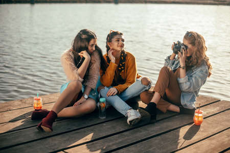 The blonde girl takes pictures of her brunette girlfriends on the old camera. They send her kisses, smile, laugh. Girl with drinks and sunglasses have fun.の写真素材