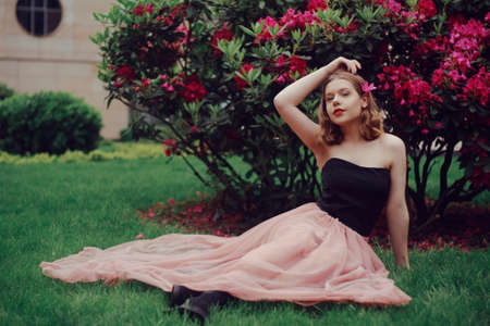 A girl in a long pink dress sits in the garden on the grass against a background of pink flowers. Short-haired blonde, fashionable princess. Flowers in her hair, looking at the camera, smiling.の写真素材