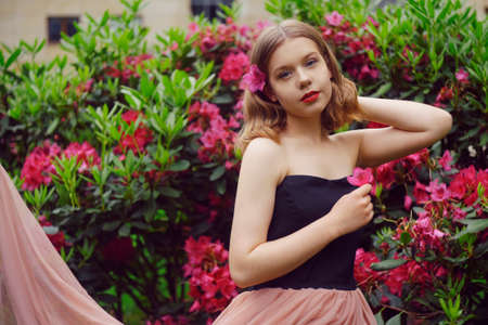 A girl in a long pink dress sits in the garden on the grass against a background of pink flowers. Short-haired blonde, fashionable princess. Flowers in her hair, looking at the camera.の写真素材