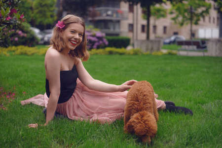 A girl in a long pink dress is sitting in the yard with her dog, a brown poodle. The girl walks with the dog, the hostess and the animal. A girl is playing with her dog.の写真素材