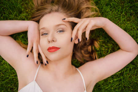 A teenager girl lies on the grass in a beautiful white dress, rests, enjoys nature. Girl with short blond hair. Beautiful and healthy skin and youth smileの写真素材