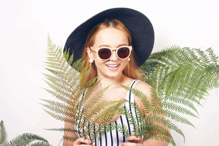 Gorgeous girl in a swimsuit, in a big black hat and sunglasses in tropical leaves rejoices in the summer. Fashion portrait. Vacation, weekend, travel, world toursの写真素材