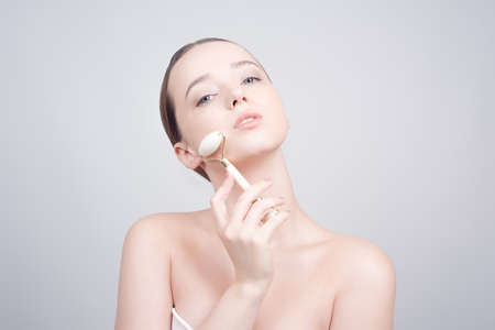 Beauty face care. Woman doing face massage with white jade facial rollers for spa skin care treatment at home. Girl model using natural massager tool portrait on whiteの写真素材