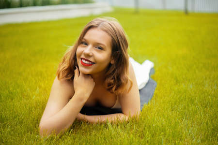 A teenager girl lies on the grass in a beautiful white dress, rests, enjoys nature. Girl with short blond hair. Beautiful and healthy skin and youth smileの写真素材