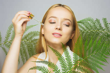 Girl on the background of tropical leaves natural cosmetics and medicines. The model holds a bottle and oil for the face and applies it to the skin. Bottle with a pipette.の写真素材