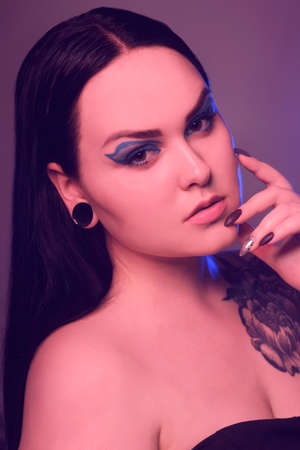 Brunette girl with a flower tattoo on her shoulder and tunnels in her ears in beautiful blue light. Woman with purple make-up, fashionable blue arrows over the eyelid.の写真素材