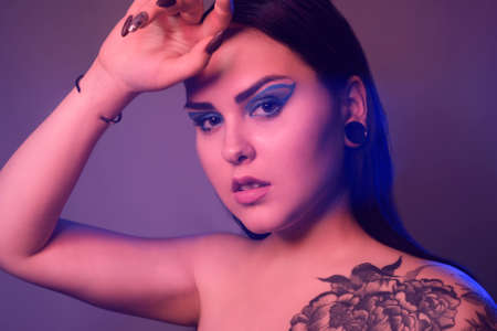 Brunette girl with a flower tattoo on her shoulder and tunnels in her ears in beautiful blue light....