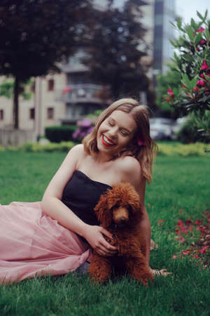 A girl in a long pink dress is sitting in the yard with her dog, a brown poodle. The girl walks with the dog, the hostess and the animal. A girl is playing with her dog.の写真素材