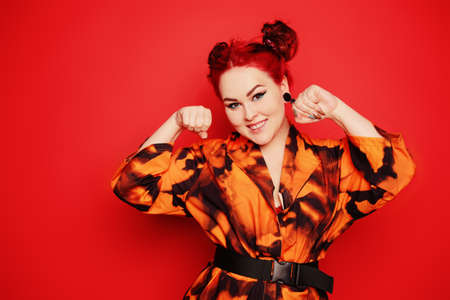 Cute girl on a red background. Red hair and tunnels in the ears, red dress and lips. Anime poses. The girl with eyeliner and two buns on her head grimaces, smiles, has fun. Japanese cultureの写真素材