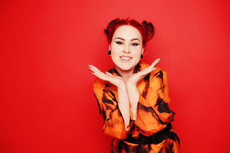 Cute girl on a red background. Red hair and tunnels in the ears, red dress and lips. Anime poses. The girl with eyeliner and two buns on her head grimaces, smiles, has fun. Japanese cultureの写真素材