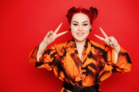Cute girl on a red background. Red hair and tunnels in the ears, red dress and lips. Anime poses. The girl with eyeliner and two buns on her head grimaces, smiles, has fun. Japanese cultureの写真素材