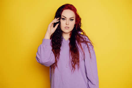 Woman in a purple sweater on a yellow background posing. A beautiful brunette with long curly hair and red roots. Eyeliner and trendy makeup. Model looking at the cameraの写真素材
