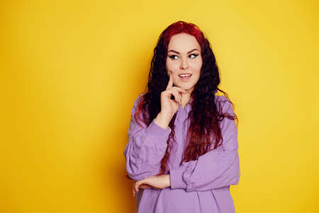 Woman in a purple sweater on a yellow background posing. A beautiful brunette with long curly hair and red roots. Model looks to the side and thinks, space for text, empty spaceの写真素材