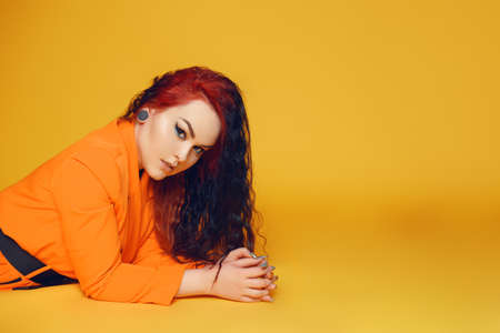 A woman with long curly hair lies on the floor against a yellow background. A girl in an orange dress with red hair and arrows in her eyes. High-fashion. Ear tunnels, earrings.の写真素材