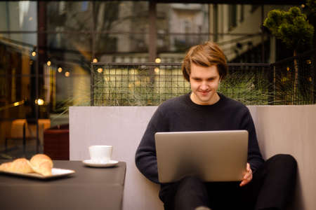 Man drinking cup of coffee in a restaurant on the terrace. Freelancer outdoor with a laptop, has breakfast with croissants. Businessman looks and dreams. Man with laptop. headphones.の写真素材