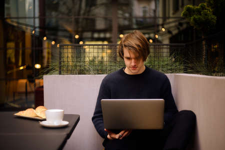 Man drinking cup of coffee in a restaurant on the terrace. Freelancer outdoor with a laptop, has breakfast with croissants. Businessman looks and dreams. Man with laptop. headphones.の写真素材